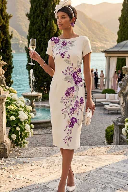 A woman stands outdoors by a lake wearing a fitted cream dress with large purple floral embroidery and holding a champagne flute. She is posed on a stone terrace with mountains, trees, and a building in the background.