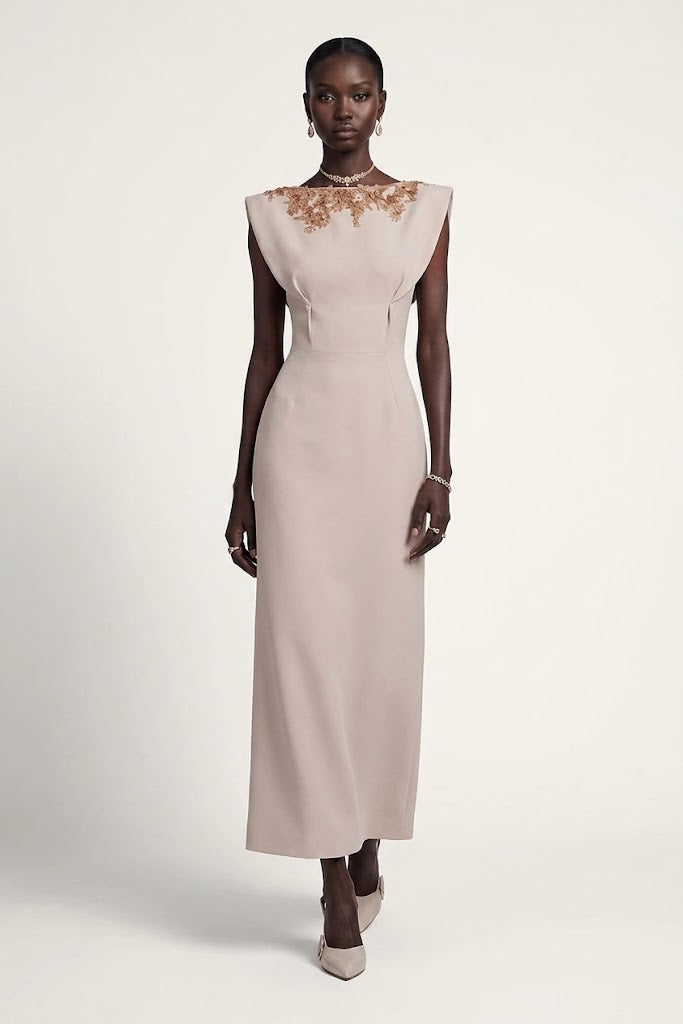 A model wearing a sleeveless, floor-length light blush pink gown with gold floral embroidery across the chest, standing against a plain off-white studio background.