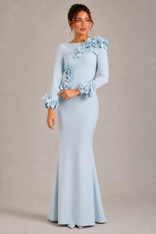 A woman modeling a floor-length light blue gown with long sleeves, ruffled floral detailing on the shoulders and cuffs, and a fitted mermaid silhouette against a plain beige studio background.