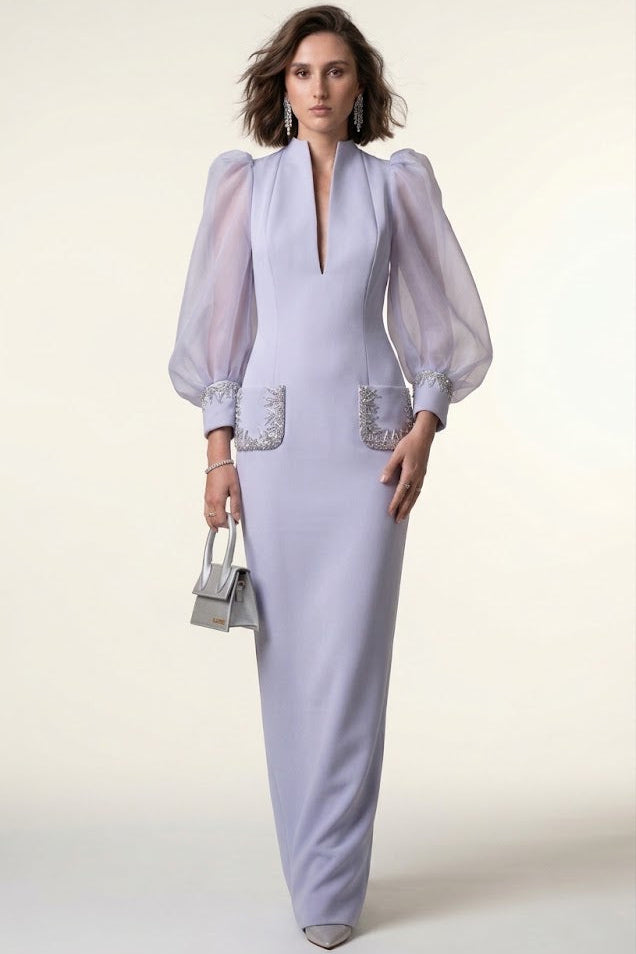 A woman models a lavender maxi dress with sheer balloon sleeves, a deep V-neckline, and silver embellished pocket details, holding a small silver handbag against a plain cream background.