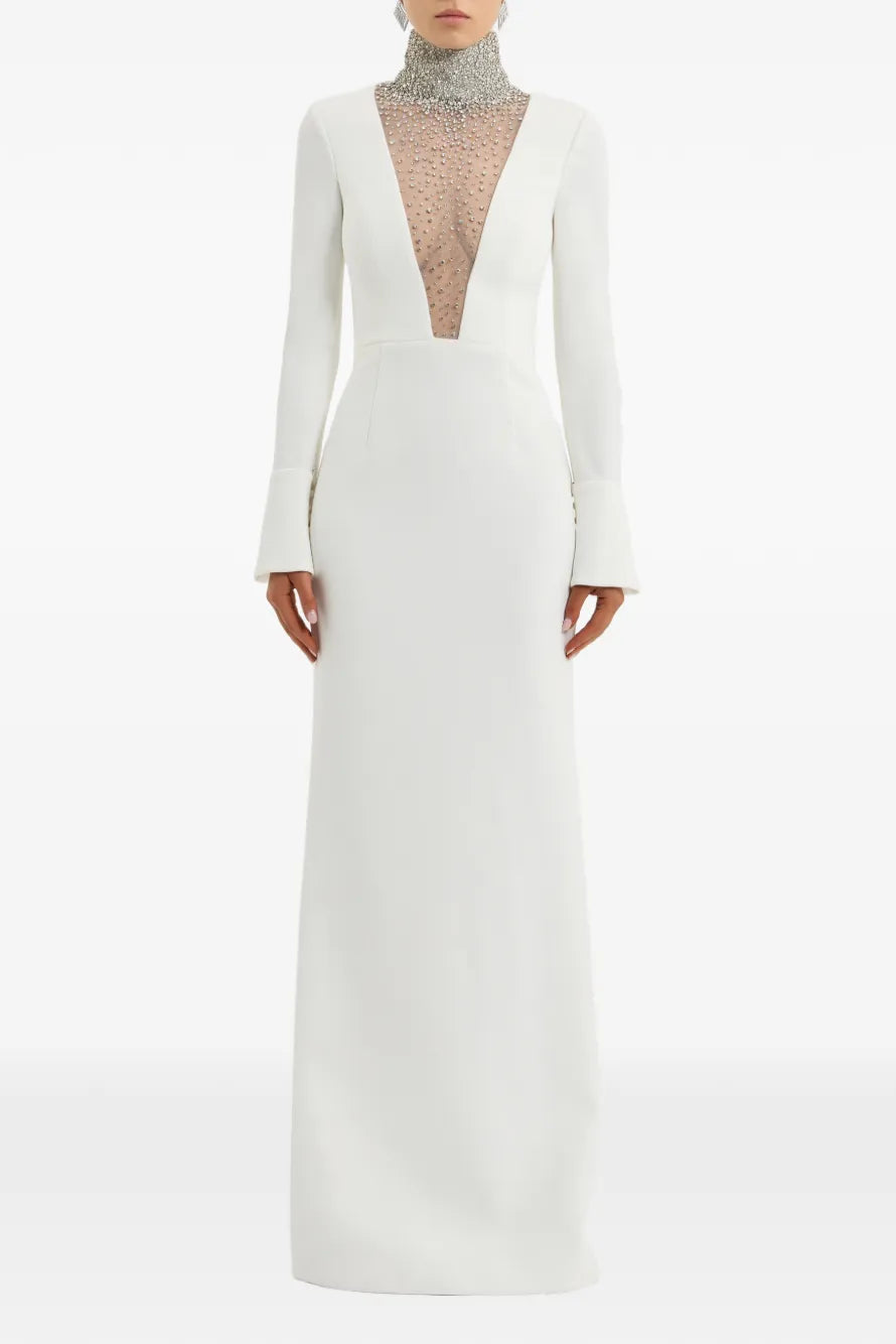 A floor-length white gown with long sleeves, a plunging V-neckline, and a sheer embellished inner bodice, shown on a model against a plain light background.