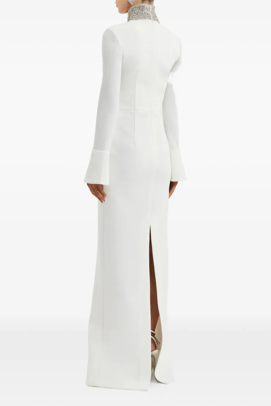 A model shown from the back wearing a floor-length white gown with long flared sleeves, a high embellished mock neck, and a front slit, photographed against a clean white studio background.