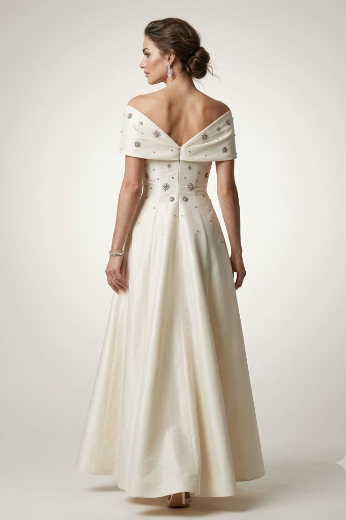 An off-the-shoulder A-line wedding dress in ivory, shown from the back on a model. The dress features delicate silver beadwork and embroidery, a deep V-back, and a full skirt, set against a plain light beige studio background.