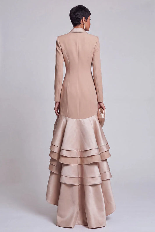 A model shown from the back wearing a formal gown with a structured beige jacket and a layered, ruffled satin skirt in a matching dusty pink-beige shade, set against a plain light gray studio background.