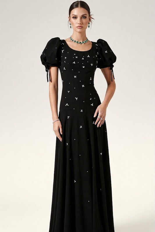 A model wearing a floor-length black evening gown with puff sleeves, tie details at the cuffs, and scattered white embroidered star-like motifs, photographed against a light gray studio background.