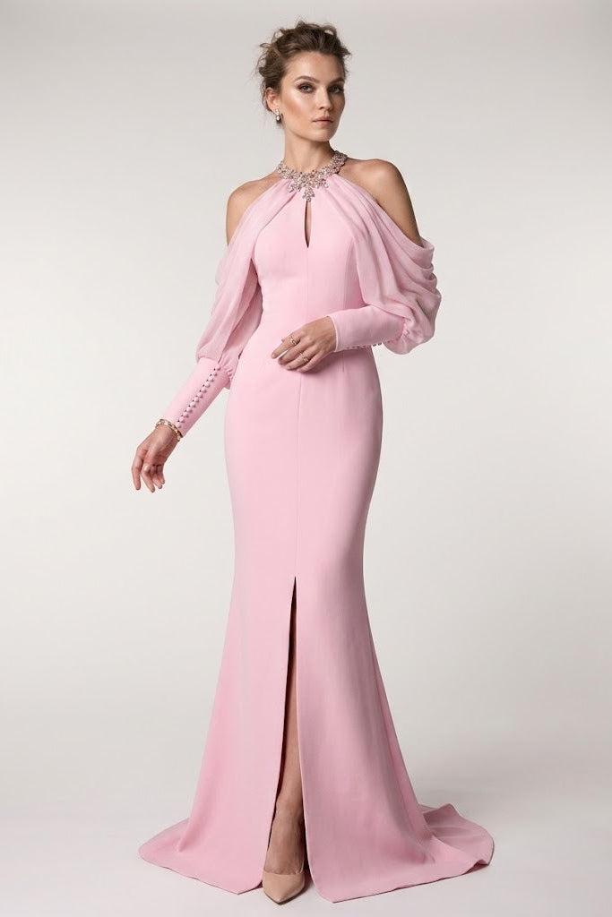 A woman models a floor-length pink evening gown with a one-shoulder design, sheer draped sleeve, and thigh-high slit, photographed against a plain light gray studio background.