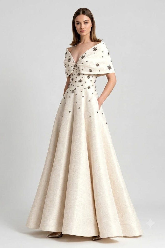 A woman modeling a floor-length off-white evening gown with a draped cape-style bodice, short sleeves, and scattered dark embroidered star motifs, photographed against a light gray studio background.