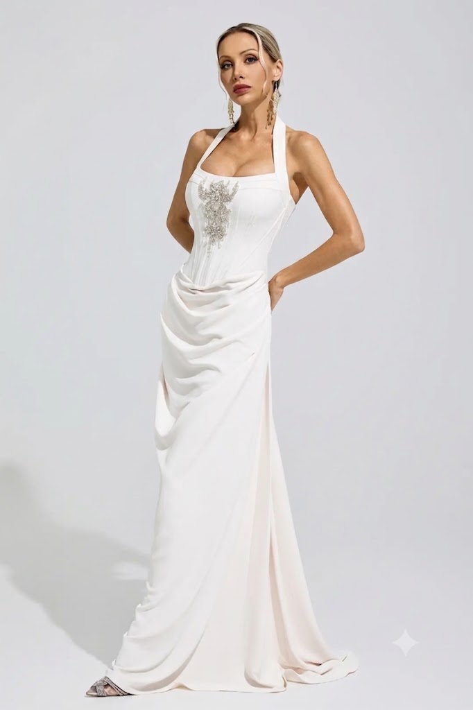 A woman modeling a floor-length white halter gown with a draped skirt and silver embellished bodice, photographed against a light gray studio background.