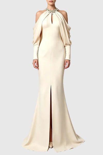 A model wearing a floor-length cream gown with a high slit, draped off-the-shoulder sleeves, and a jeweled collar detail, set against a solid light gray background.