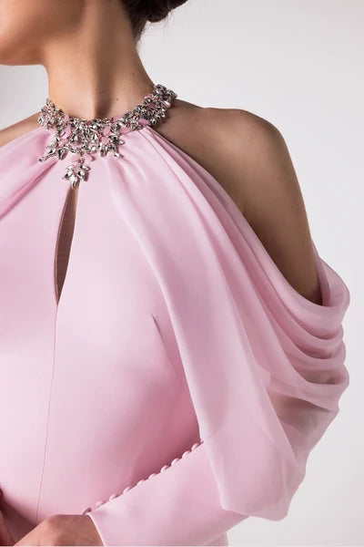 A close-up of a model wearing a light pink halter-neck gown with a high-key embellished neckline and draped sheer sleeves against a plain light background.