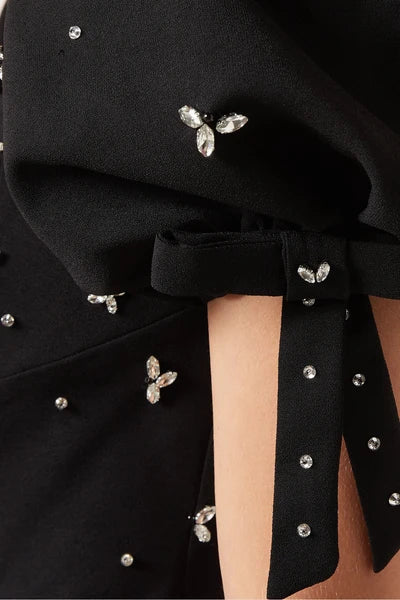 Close-up of a black dress with crystal-embellished bow details and rhinestone accents, shown on a model.