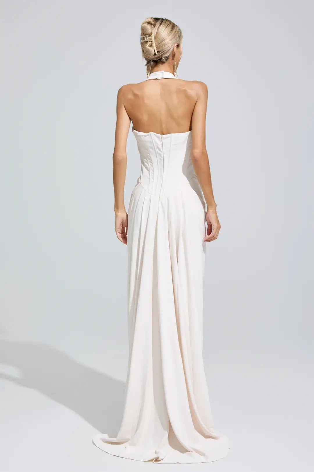 A woman shown from the back wearing a strapless halter-neck floor-length gown in a warm off-white or cream tone, photographed against a light gray studio background.