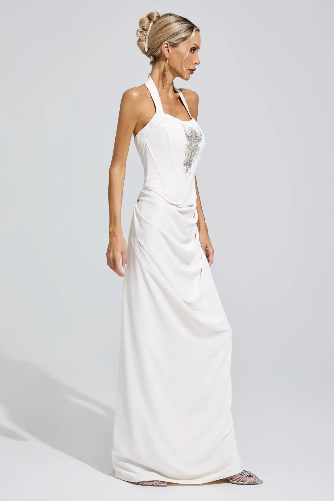 A woman modeling a white halter-neck floor-length gown with a structured bodice, draped skirt, and silver embellishment on the chest, photographed against a light gray studio background.