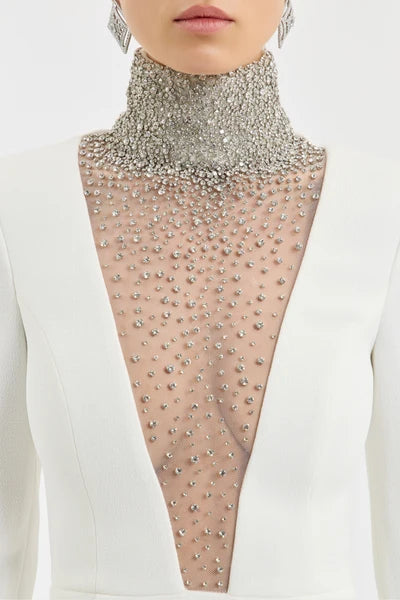 A close-up of a model wearing a white structured jacket over a sheer mesh top heavily embellished with silver rhinestones, featuring a high silver rhinestone collar.