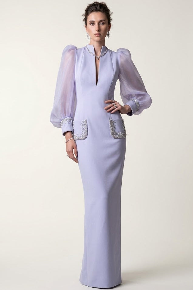 A model wears a lavender floor-length gown with sheer balloon sleeves, a split neckline, and silver embellished pockets, set against a plain light beige studio background.