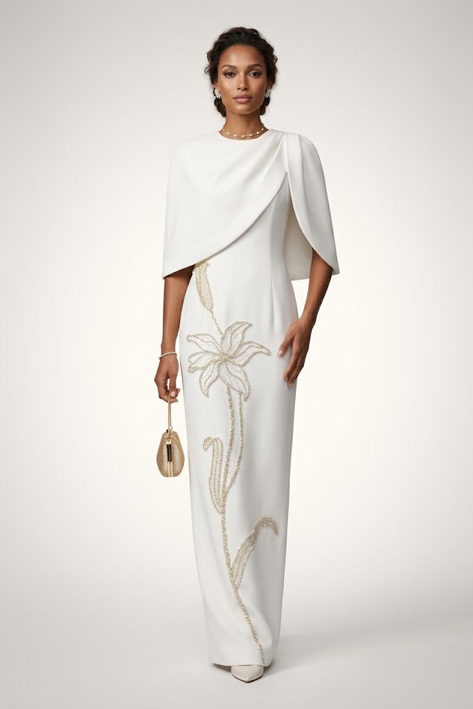 A woman models a floor-length white gown with draped cape-like sleeves and a vertical floral embellishment in a light gold/beige tone. She is standing against a plain light gray studio background and holding a small metallic clutch.