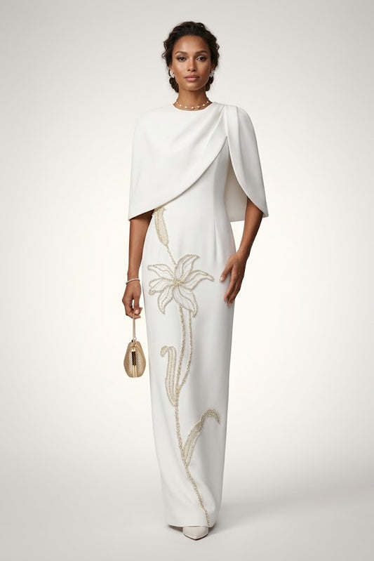A woman models a floor-length white gown with draped cape-like sleeves and a vertical floral embellishment in a light gold/beige tone. She is standing against a plain light gray studio background and holding a small metallic clutch.