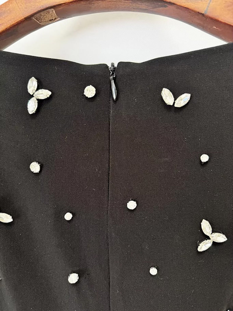 Close-up of a black garment with a central zipper and scattered rhinestone embellishments, including small white dots and silver-tone leaf clusters, shown against a white background with part of a wooden hanger visible at the top.