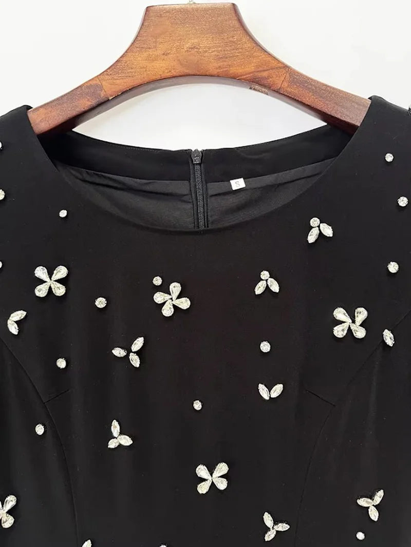 A close-up of a black sleeveless top or dress with scattered white floral and dot embellishments, hanging on a wooden hanger against a plain light background.