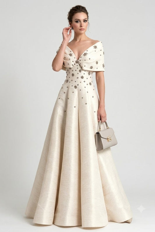 A woman models a floor-length off-the-shoulder gown in warm ivory or cream satin with scattered metallic gold floral embroidery. She is holding a small structured beige handbag against a plain light gray studio background.