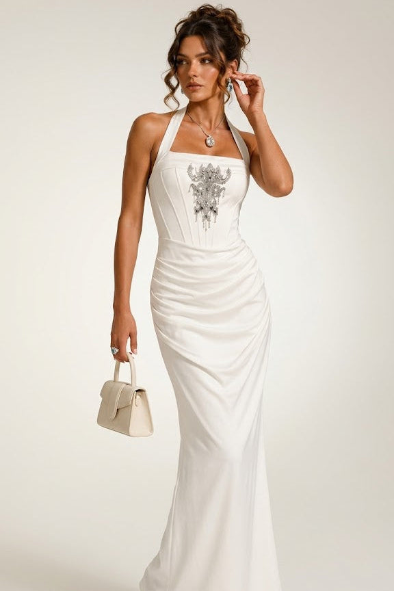 A woman models a white halter-neck maxi dress with a silver graphic print on the bodice, holding a small beige handbag against a plain light background.