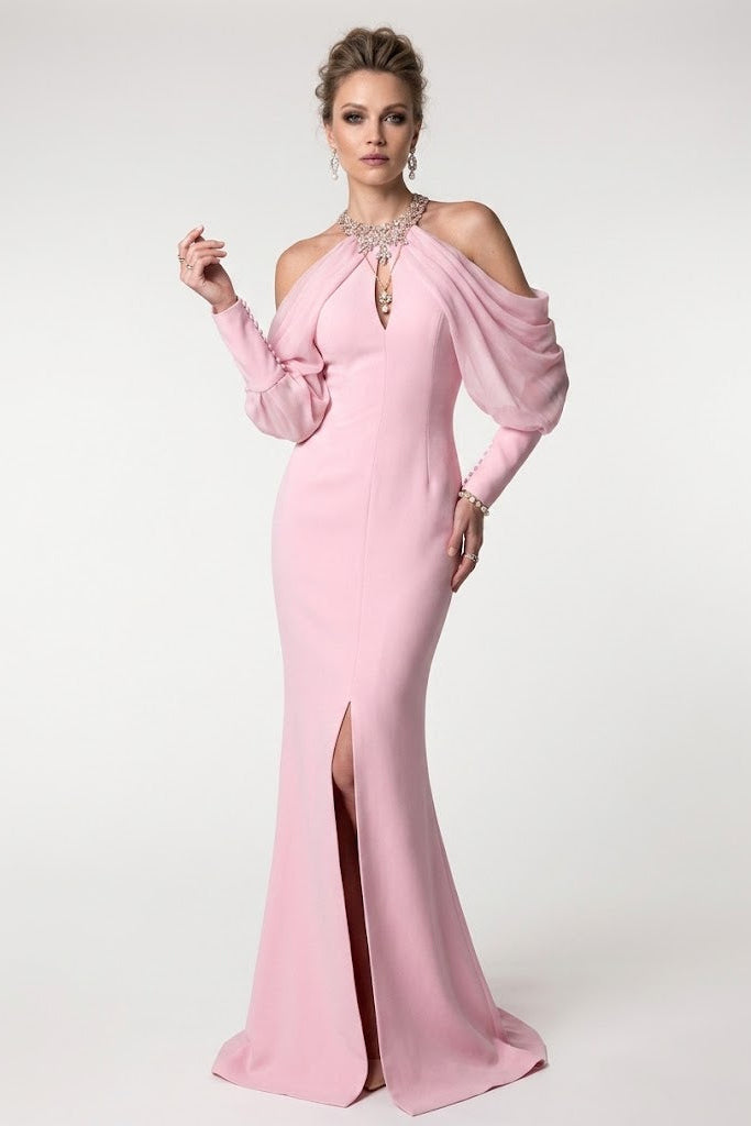 A woman modeling a light pink cold-shoulder evening gown with a high thigh slit, long draped sleeves, and a beaded neckline, set against a plain light gray studio background.